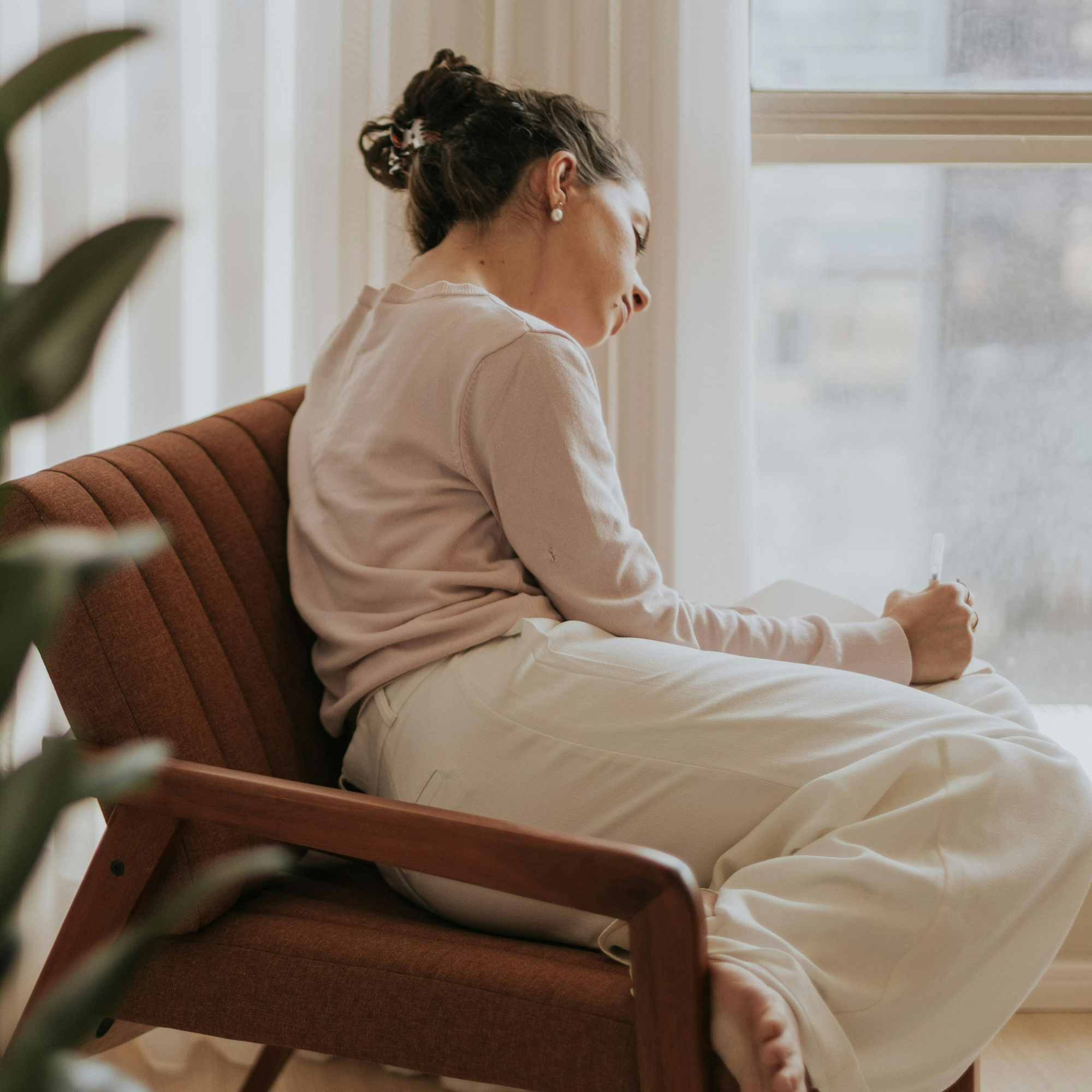 Woman sitting in a brown armchair by a window, writing in a notebook